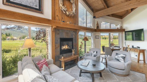 Cozy living room with a fireplace, large windows, and a view of greenery, featuring comfortable seating and wooden accents.